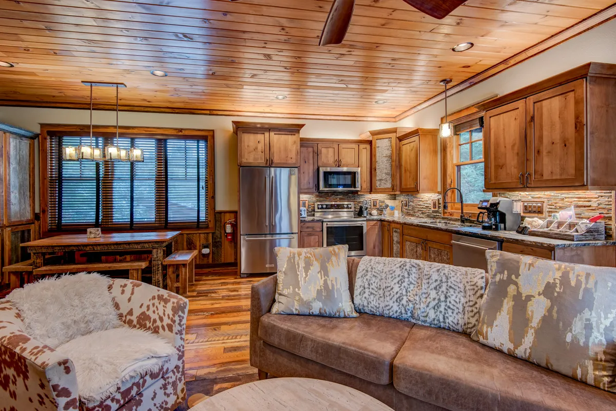 An open-plan view showing a comfortable sofa, a dining table ready for group meals, and a kitchen island—all framed by large windows that fill the space with natural light. The setup encourages easy flow between relaxing, dining, and cooking.
