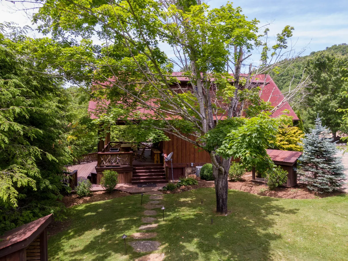 This image features a rustic wooden house with a red roof nestled among lush greenery, with a garden path leading up to the porch. The mature tree in the front yard and surrounding landscape evoke a sense of tranquility and year-round natural beauty. The porch and outdoor area hint at gatherings and outdoor activities in warmer seasons.