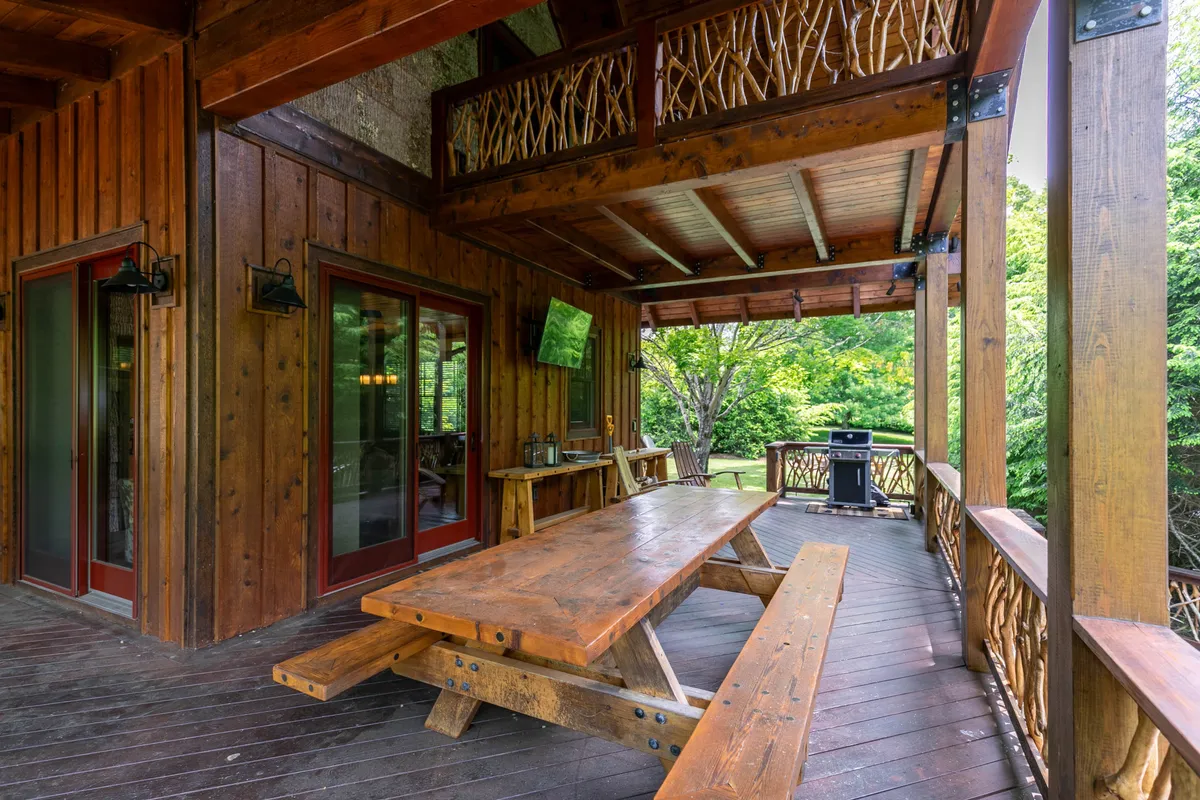 A welcoming wooden balcony featuring a dining table, balcony railing, grill, and large windows connecting back to the home’s interior. The scene is set for outdoor dining, barbecues, or sunset celebrations against a backdrop of natural scenery.