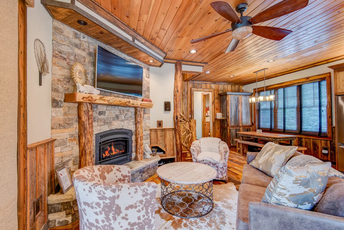 A warm, welcoming living room centered around a striking stone fireplace, with comfortable seating arranged for conversation and relaxation. The wooden ceiling and round coffee table enhance the cozy, rustic mountain atmosphere. Large windows suggest tranquil views just outside, and the space flows naturally towards a dining area.