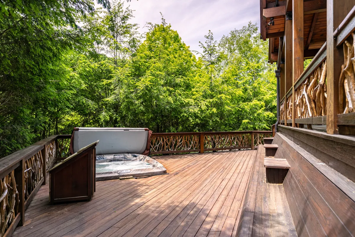 A spacious wooden deck set amidst lush greenery, featuring a state-of-the-art hot tub and a decorative railing. The deck offers a tranquil spot for relaxation, yoga, or social gatherings, with the hot tub perfectly positioned to soak up the surrounding natural beauty.