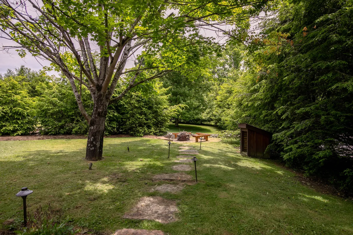A large tree shades a rustic fire pit area with a wooden table and a meandering pathway, set within a green, open space. The environment feels natural and playful, ideal for outdoor games, picnics, and stargazing as a family.
