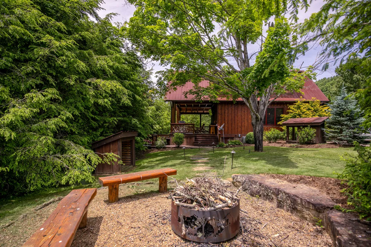 A cozy outdoor fire pit area surrounded by rustic wooden benches, nestled in a lush green setting near a cabin structure. The space invites gatherings under the stars, creating a warm atmosphere for storytelling and s’mores.