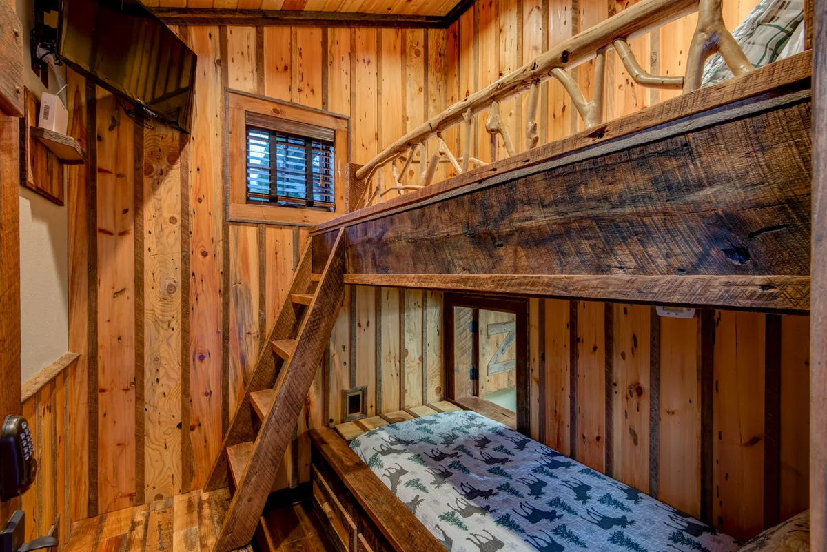 This image displays a cozy bunk room with bunk beds nestled against textured wooden walls. A small window lets in natural light, and the space is framed by a rustic wooden staircase, evoking the sense of a hidden nook. The environment feels playful and intimate, ideal for children or anyone seeking a secret hideaway.