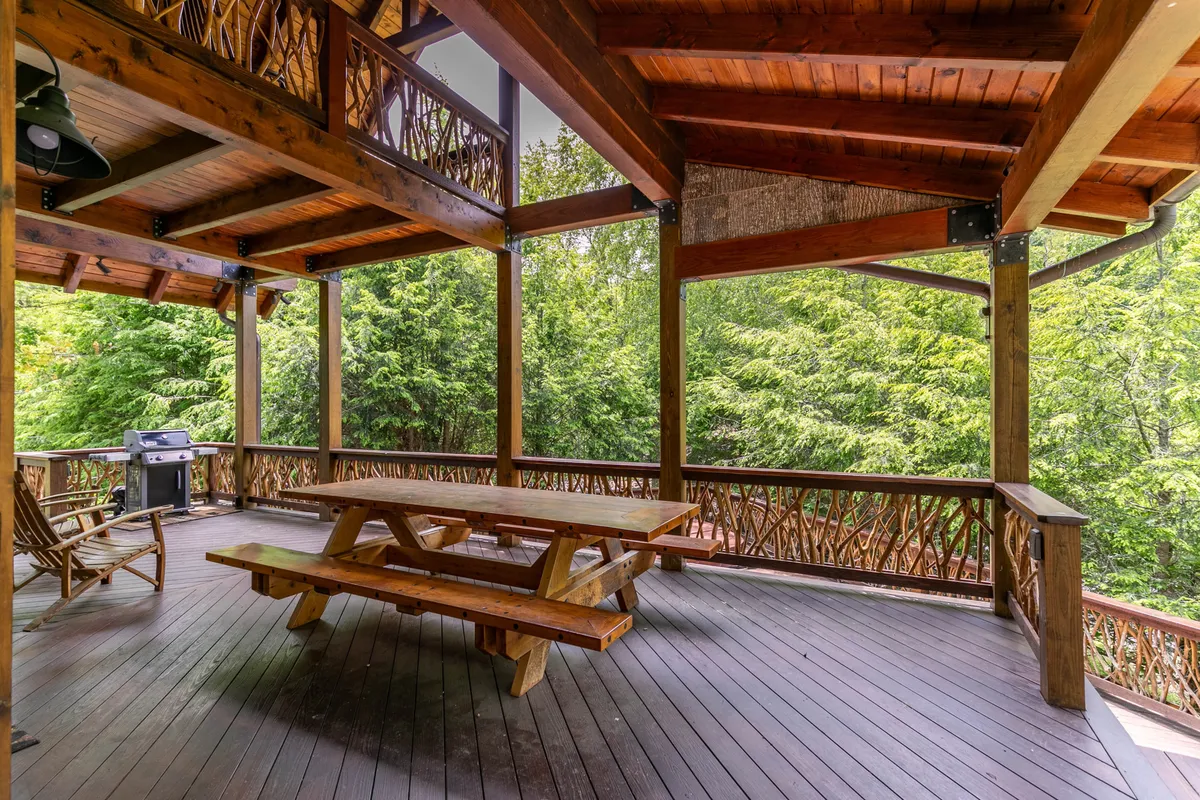 This image depicts a welcoming outdoor setting with a wooden picnic table, classic rocking chairs, a grill, and lush greenery all around. The scene suggests a communal, family-friendly area perfect for outdoor meals, games, or simply relaxing and enjoying nature together.