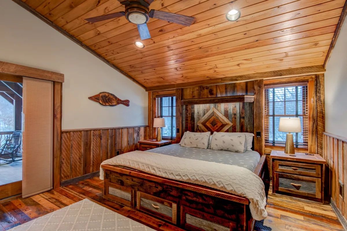 This image showcases a warmly inviting bedroom with a wooden bed frame, decorative headboard, cozy lamps on the nightstands, wood-paneled walls, and a sliding door—likely leading to a private deck. Natural light subtly fills the space, highlighting the plush bedding and tranquil atmosphere.