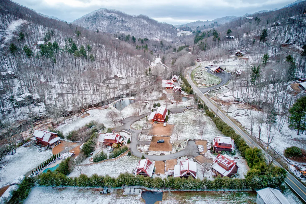 This expansive exterior shot captures snow-covered cabins, a winding road, mountain backdrop, and a tranquil pond surrounded by greenery. The scene embodies both adventure and peacefulness in the heart of the mountains, suggesting opportunities for hiking, photography, and enjoying nature's seasonal changes.