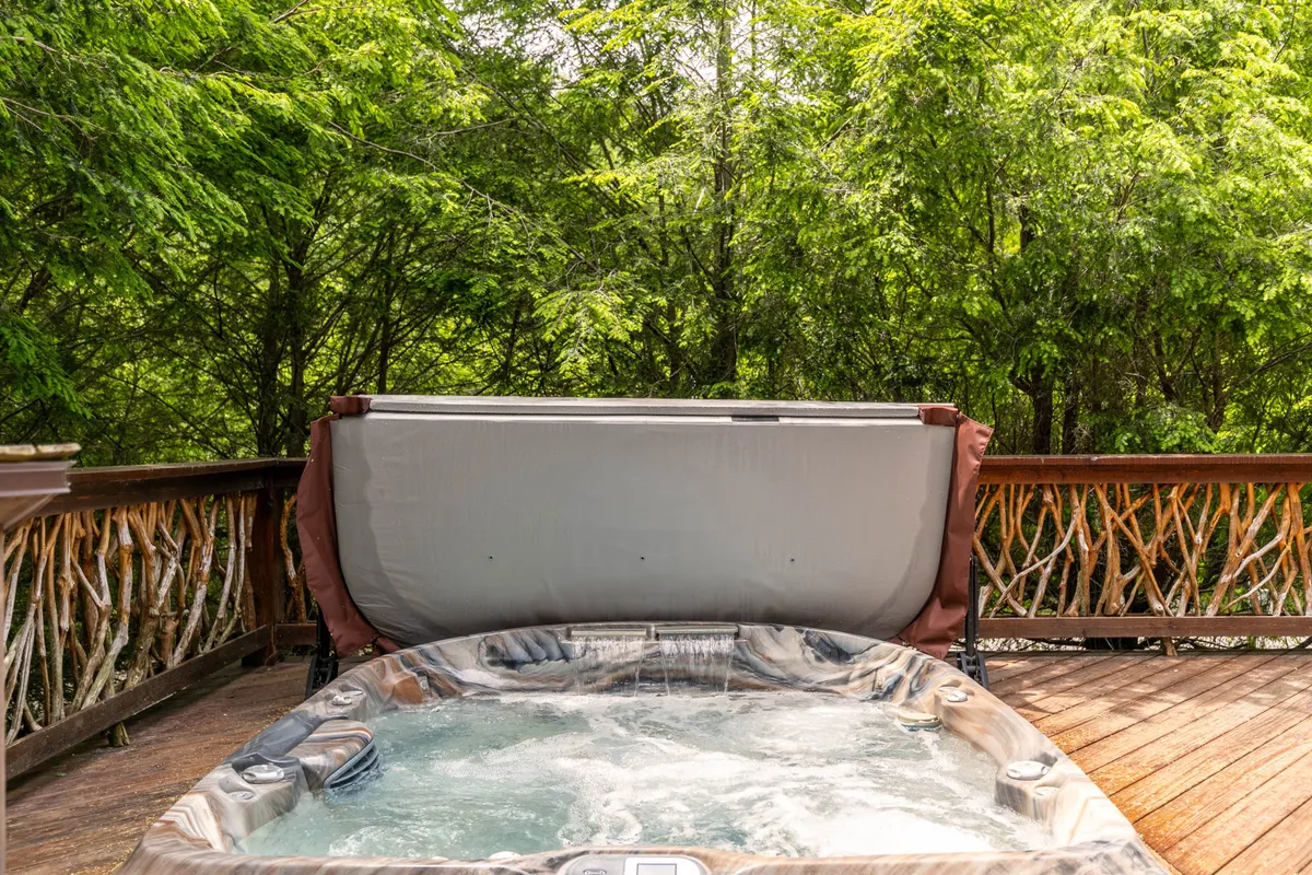 This image showcases a hot tub set on a wooden deck, surrounded by lush greenery. The tranquil setting is perfect for relaxing while overlooking the river, with soft natural light suggesting the peacefulness of sunset or twilight.