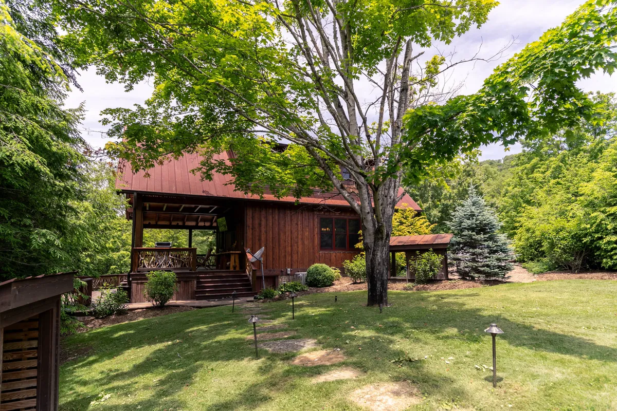 A cozy wooden house set amid a landscaped garden, with a large tree providing shade over a patio area. The setting is peaceful and inviting, ideal for outdoor dining, family gatherings, or simply soaking up the tranquil environment. The house's charming exterior emphasizes the private, standalone cabin feel.