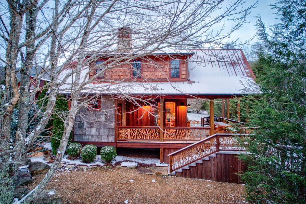 A charming wooden porch with a shingled roof and stone chimney sits amid a snowy landscape. The snow-covered ground, cozy home, and hints of warmth from inside evoke the comforts of winter and the promise of snow play, followed by relaxation in a welcoming retreat.