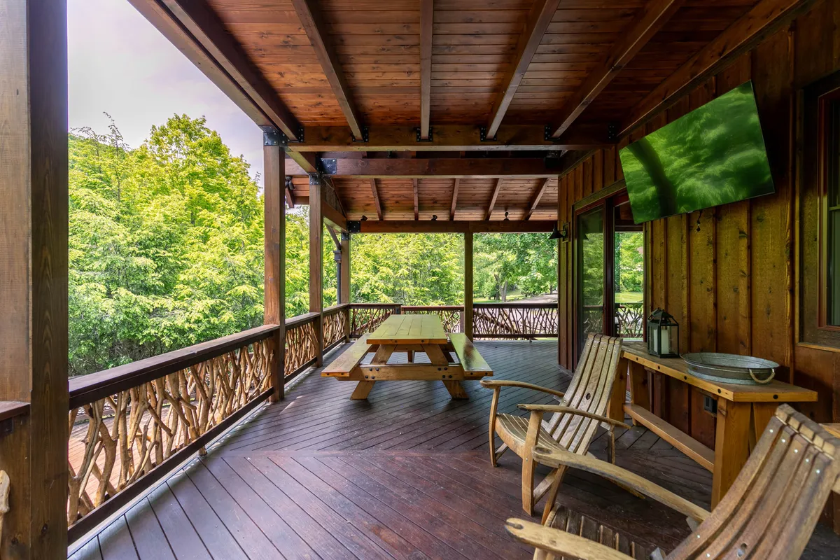 This exterior shot features a wrap-around porch with Adirondack chairs, a wooden table for outdoor dining, a mounted television, and vibrant greenery. The space is casual and welcoming, inviting families to relax, dine, or play games outdoors while staying connected to nature.